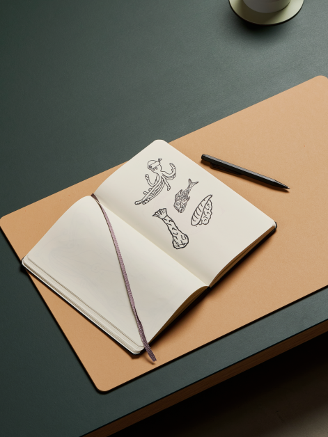 Areal view of a linoleum table with a Faust medium-format PAD Mat in 4001 Clay linoleum with sketchbook and a pen, creating a comfortable writing surface