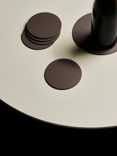 View of a round linoleum table featuring seven linoleum PAD coasters in Mauve shade, with a bottle of wine placed on the largest coaster and a glass ring mark visible on one small coaster