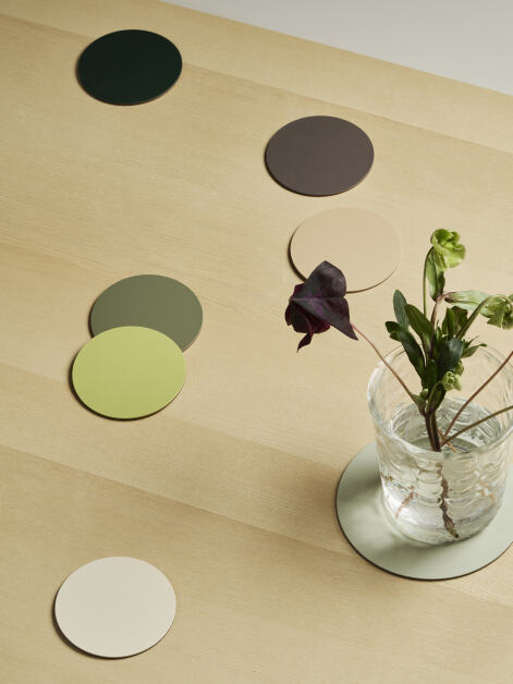 View of an oak veneer table featuring seven linoleum PAD coasters in forest-toned colors, with a transparent glass vase containing water and flowers placed on the largest coaster