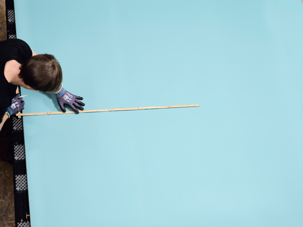 Tischplattenzuschnitt mit Linoleum in Hellblau – eine Hand fixiert einen Zollstock auf der Platte, Arbeitssituation bei der Maßaufnahme.