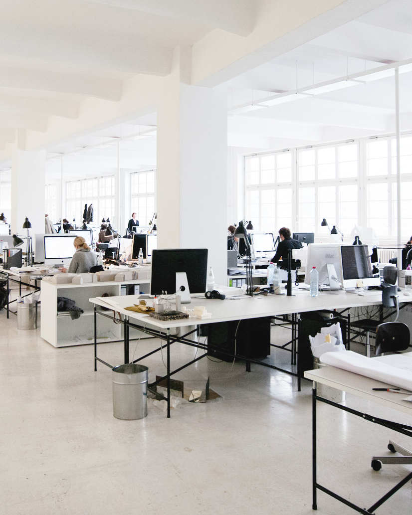 E2 desks with light gray linoleum tops in a large architecture office, used as workstations with computers and models.
