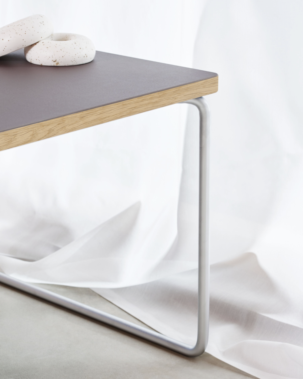 Low & Lower side table by Faust with linoleum surface in Mauve, oak edge and silver frame, in front of a white curtain.