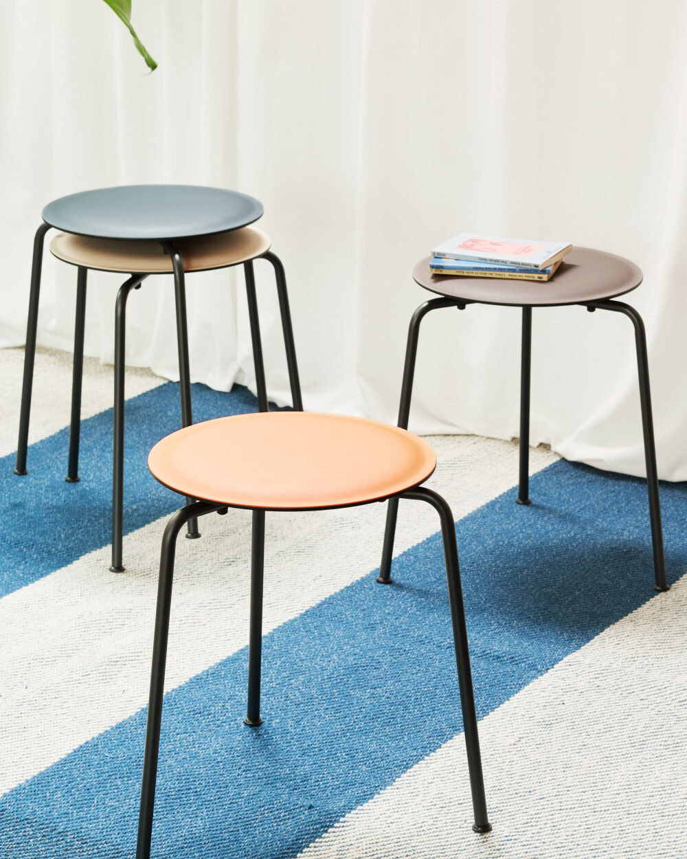 RAVIOLI Hocker mit Linoleum-Sitzflächen in verschiedenen Farben auf blau-weißem Teppich vor hellen Vorhängen.