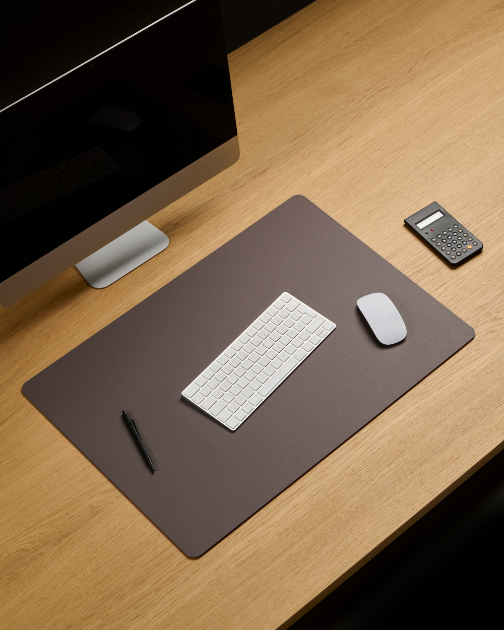 Desk with espresso-coloured linoleum PAD mat, keyboard, mouse and pen on top; monitor in the background.